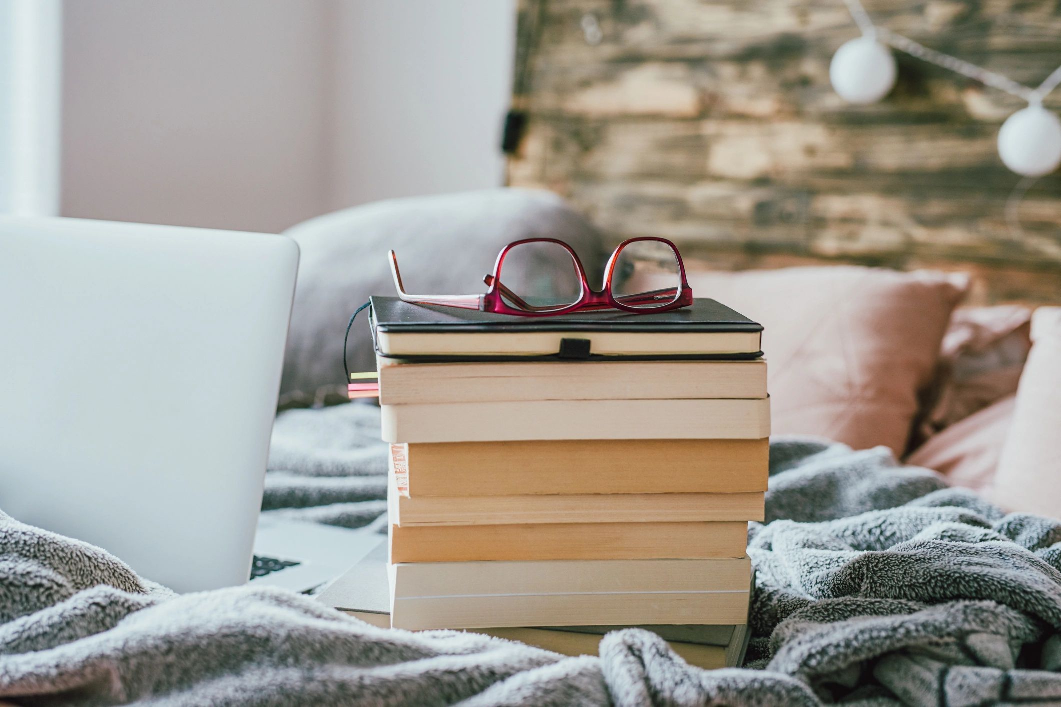 Stack of books on a bed with glasses and a laptop